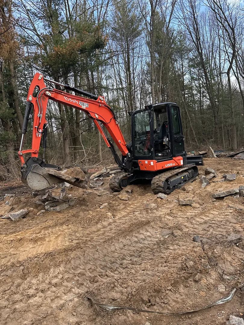 Compact excavator moving rocks in a forested area, surrounded by bare trees and earth.