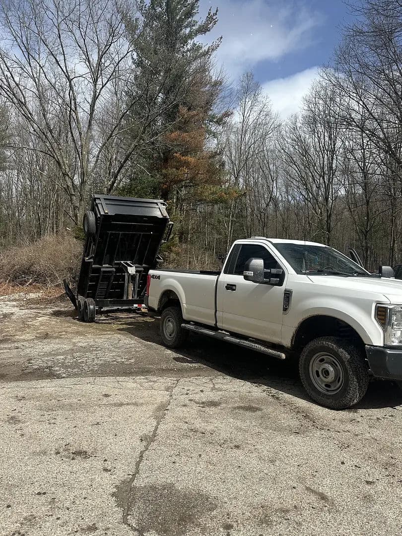 White pickup truck unloading a trailer in a forested area on a cloudy day.