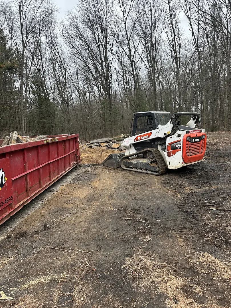 Bobcat skid steer near a debris dumpster in a wooded area, surrounded by logs and clearing equipment.