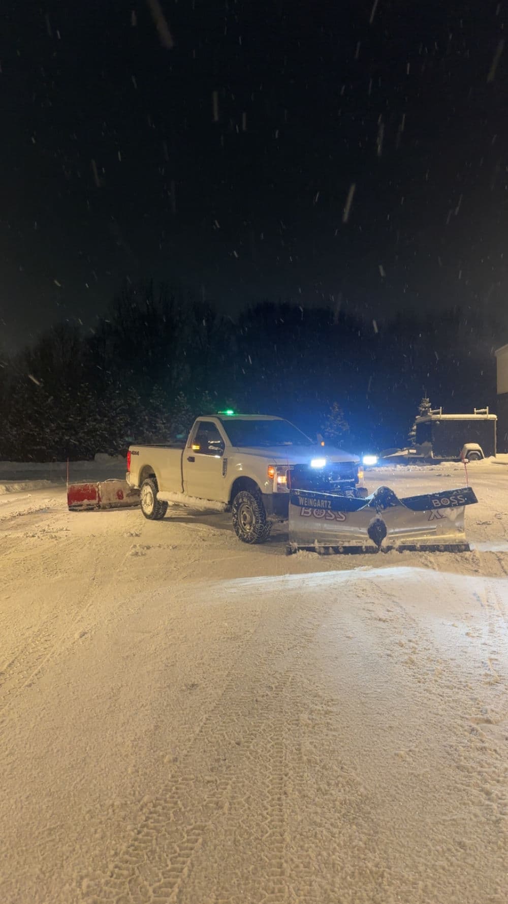 Snow-covered truck with plow clears driveway during nighttime snowstorm.