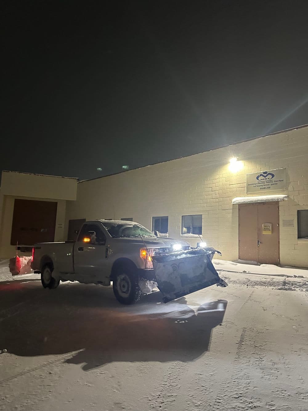 Pickup truck with snow plow at night in front of a commercial building, illuminated by lights.