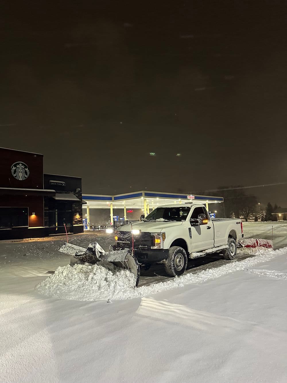 White truck with plow clearing snow at night near a gas station and Starbucks.