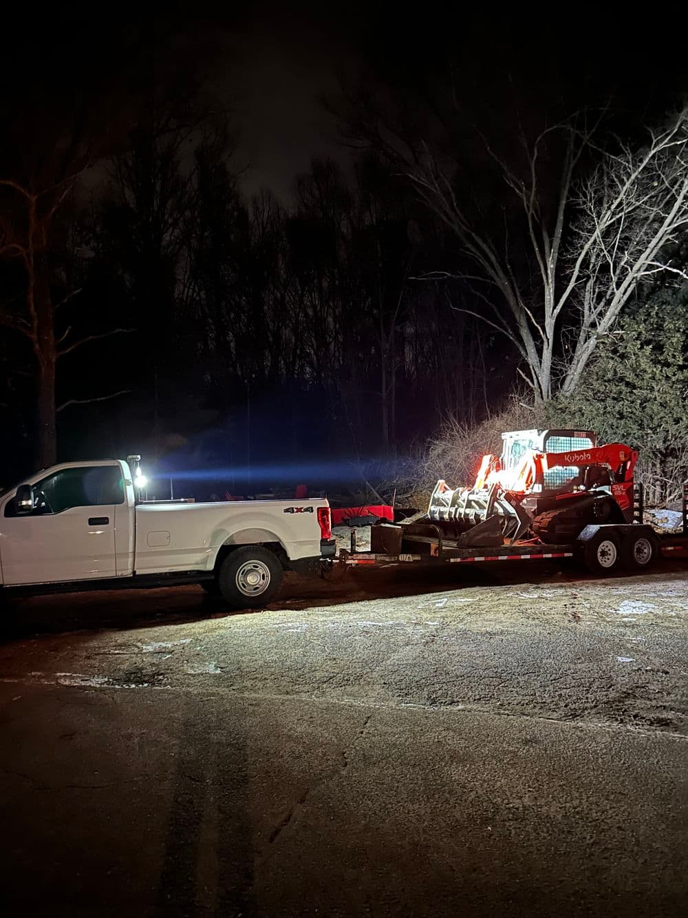 White pickup truck towing a red tractor at night with illuminated surroundings.