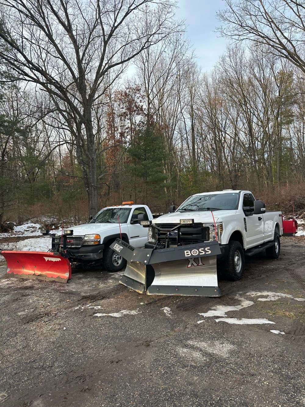Two pickup trucks with snow plows parked in a wooded area during winter.