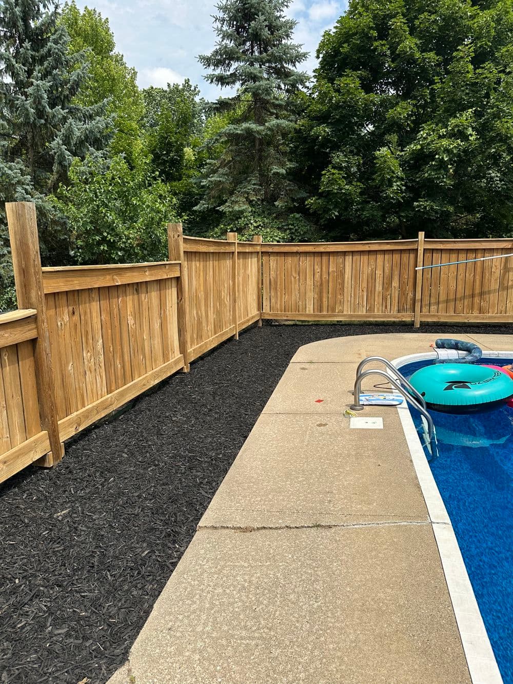 Wooden fence surrounding a pool area with blue water and green trees in the background.