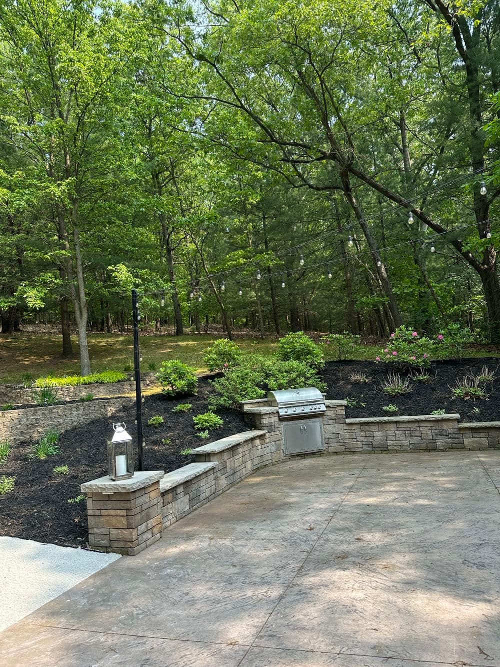 Outdoor patio with stone seating, grill, and surrounded by lush greenery and trees.