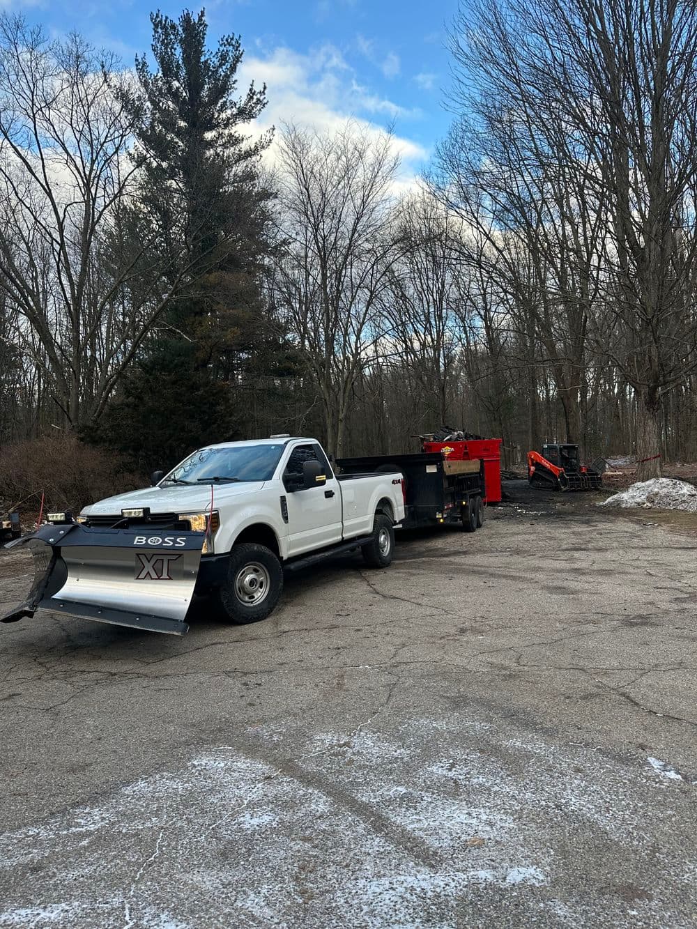 White truck with plow and trailer in wooded area, surrounded by trees and snow.
