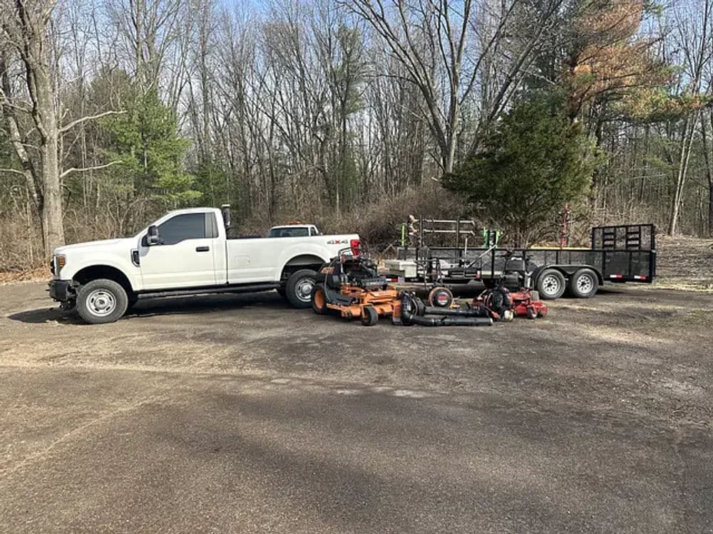 White pickup truck with lawn care equipment and trailer parked in a wooded area.
